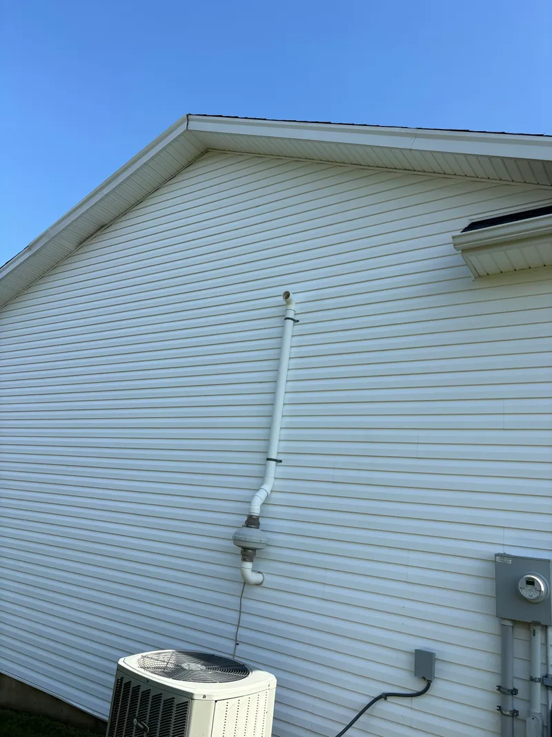 After cleaning: white vinyl siding free of staining, with a vent pipe and electrical box on the wall and an AC unit below, under a clear blue sky.