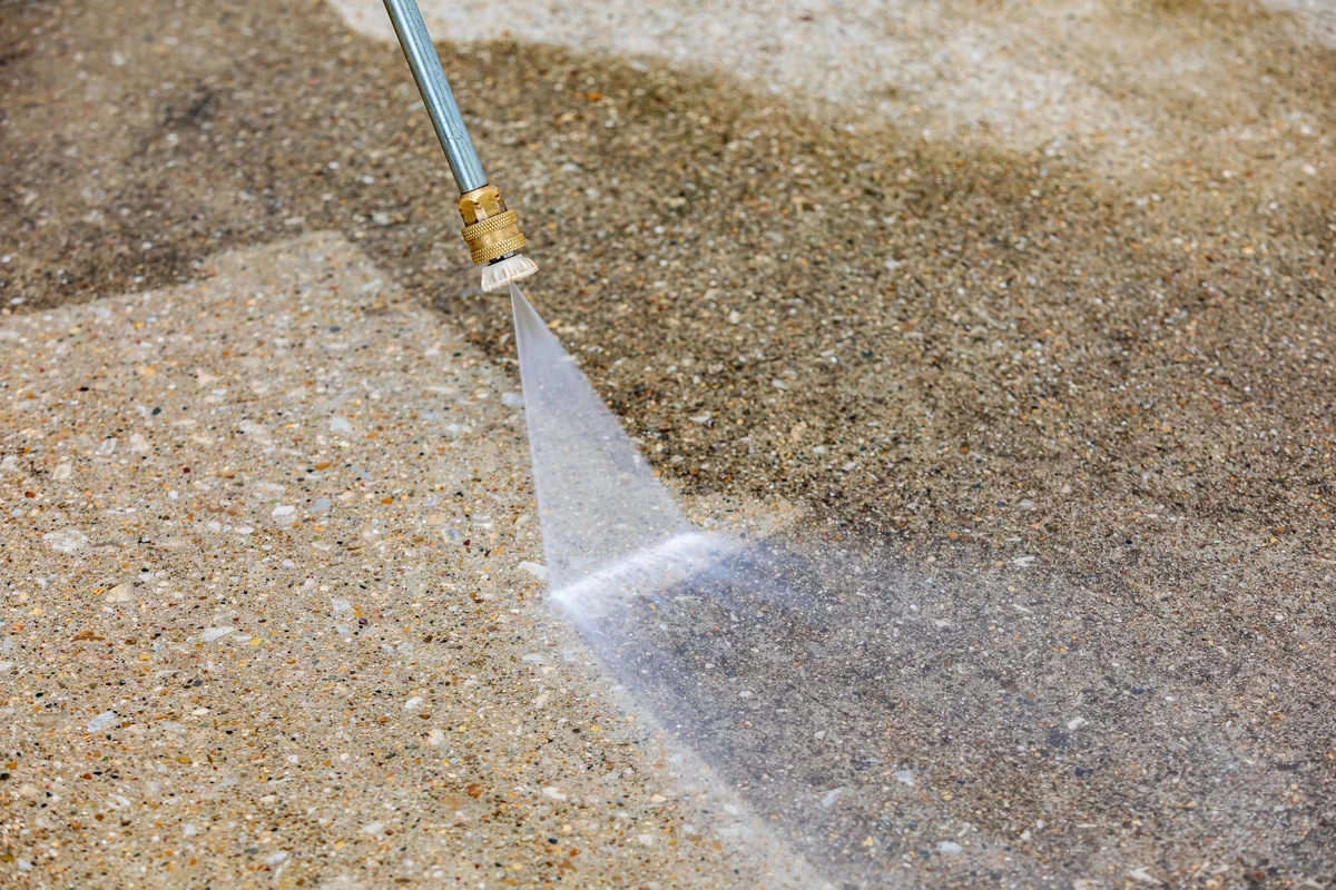 Pressure washing wand cutting a clean wedge across dirty exposed aggregate concrete