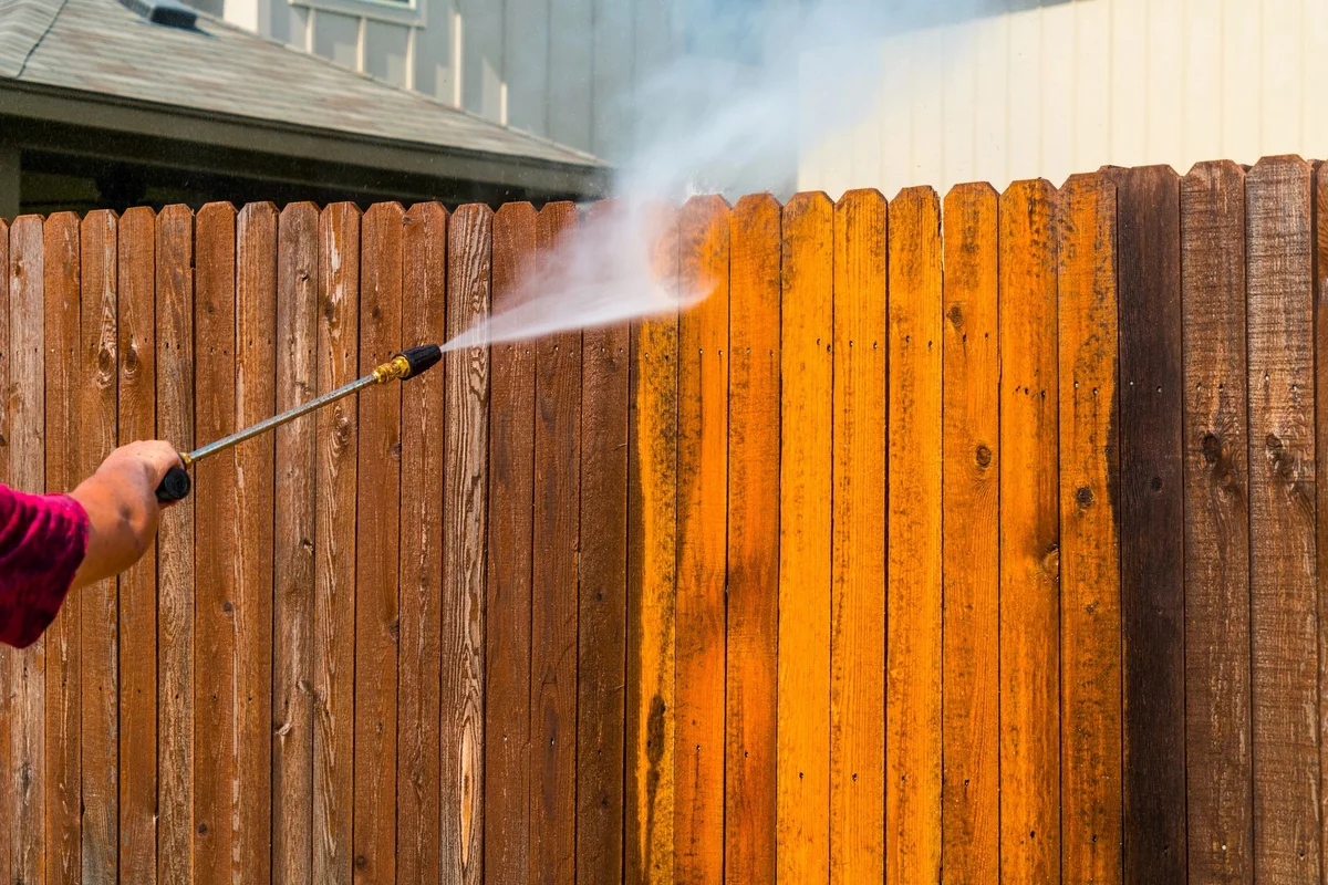 Wand spraying a wood privacy fence, leaving clean gold wood beside dark weathered boards