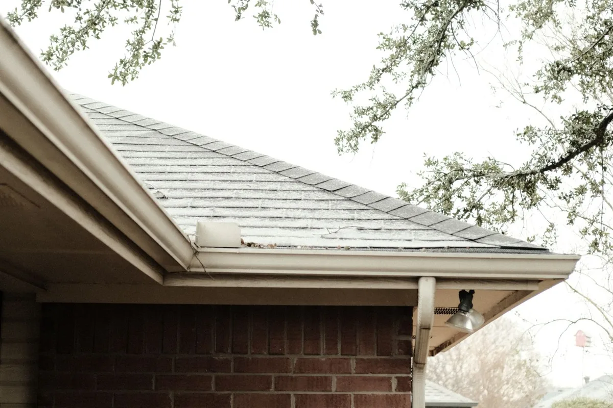 A cream gutter running the length of the eaves on a red brick house, with the downspout at the corner and the shingle roof above, seen from the ground.