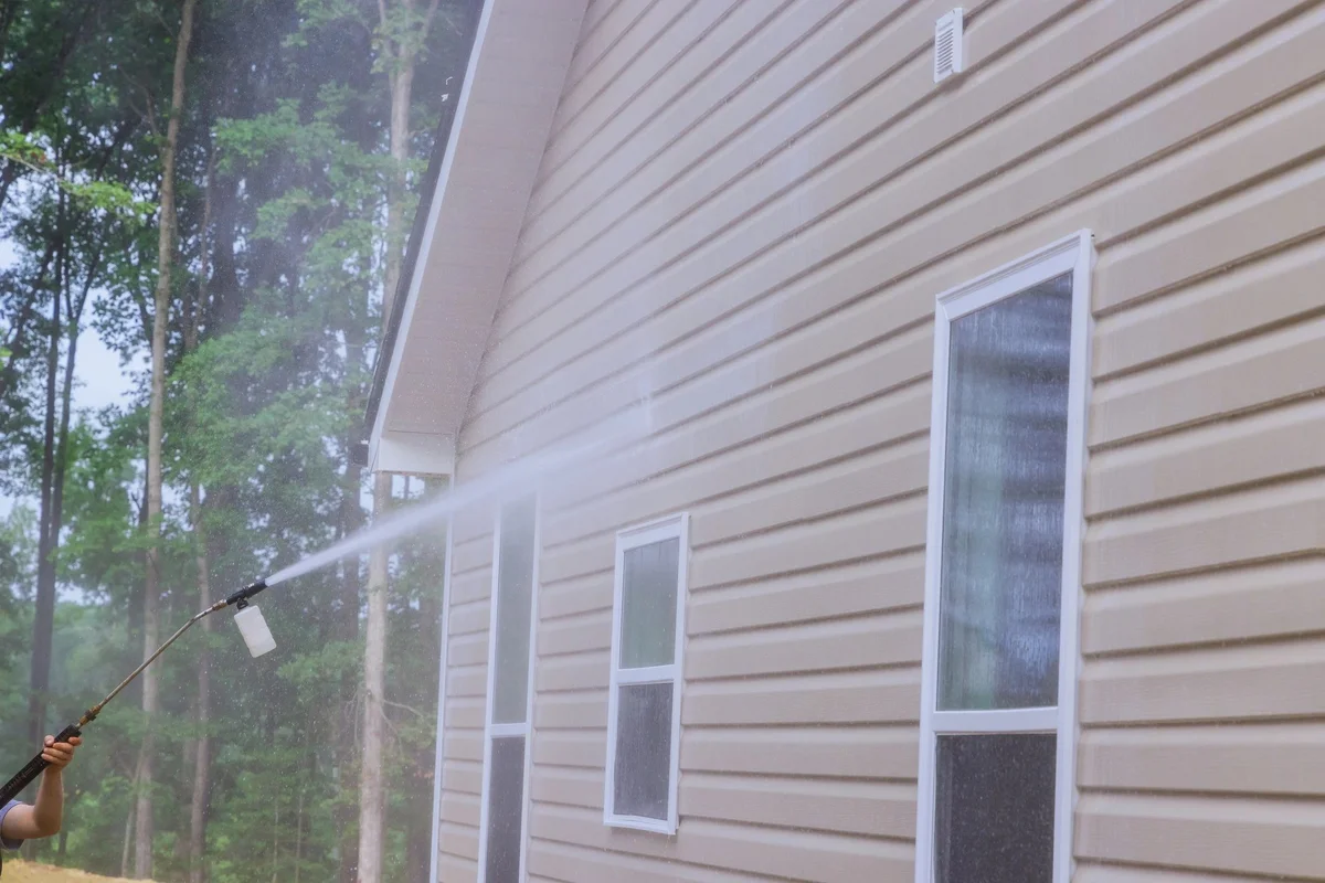 Spray wand aiming a fan of water at tan vinyl siding near the gable peak of a two-story house