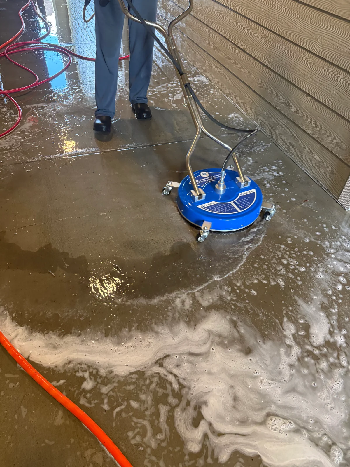 Person in dark shoes guiding a blue surface cleaner across a wet concrete floor, with soap foam and hoses nearby