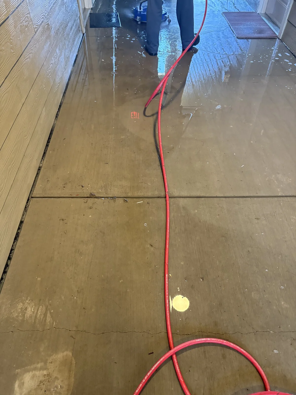 Wet concrete breezeway floor with a red hose running toward a person in dark pants and a blue surface cleaner at the far end