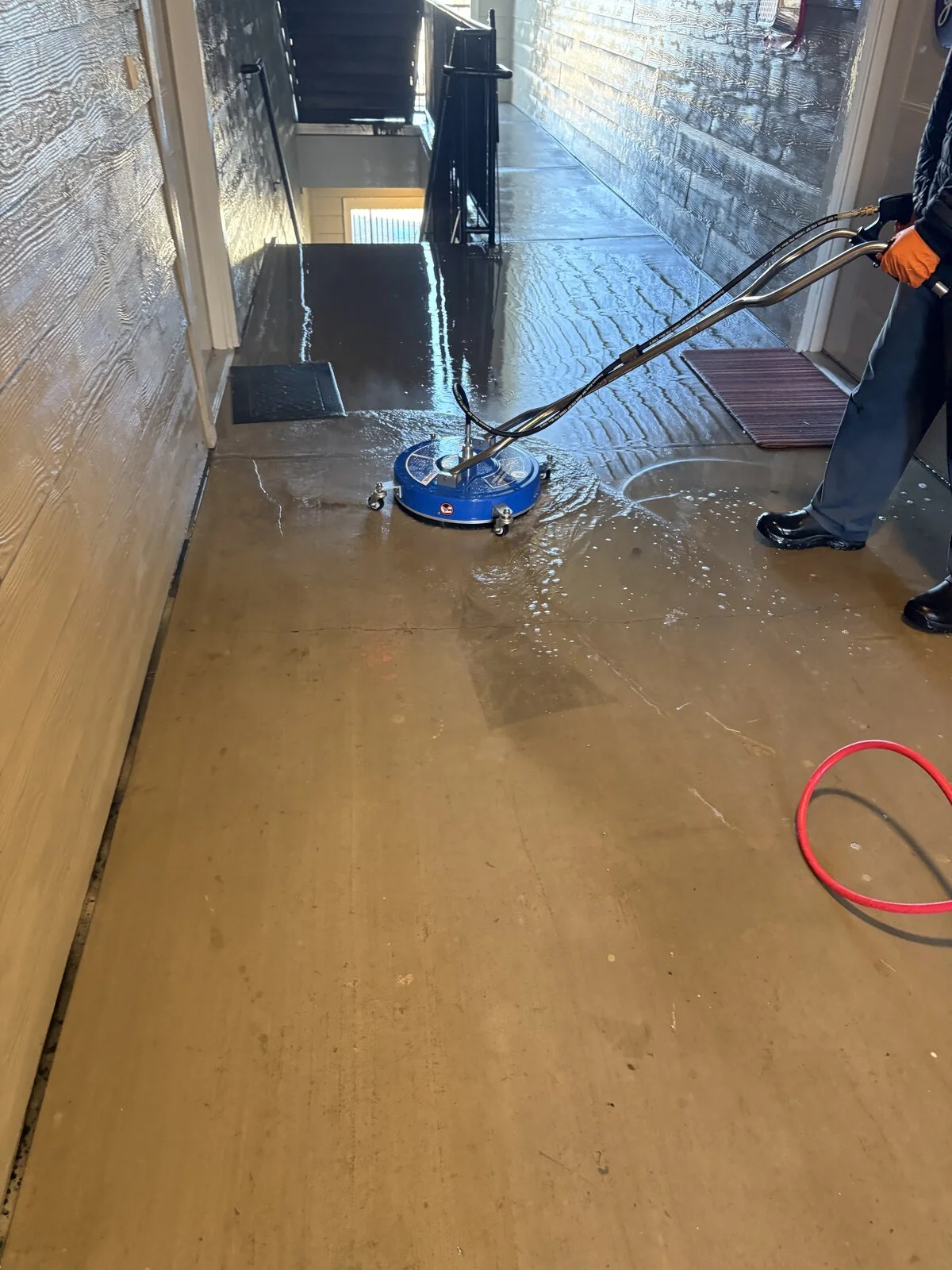 Person in orange gloves guiding a blue surface cleaner across a wet concrete floor in a long breezeway with textured siding walls