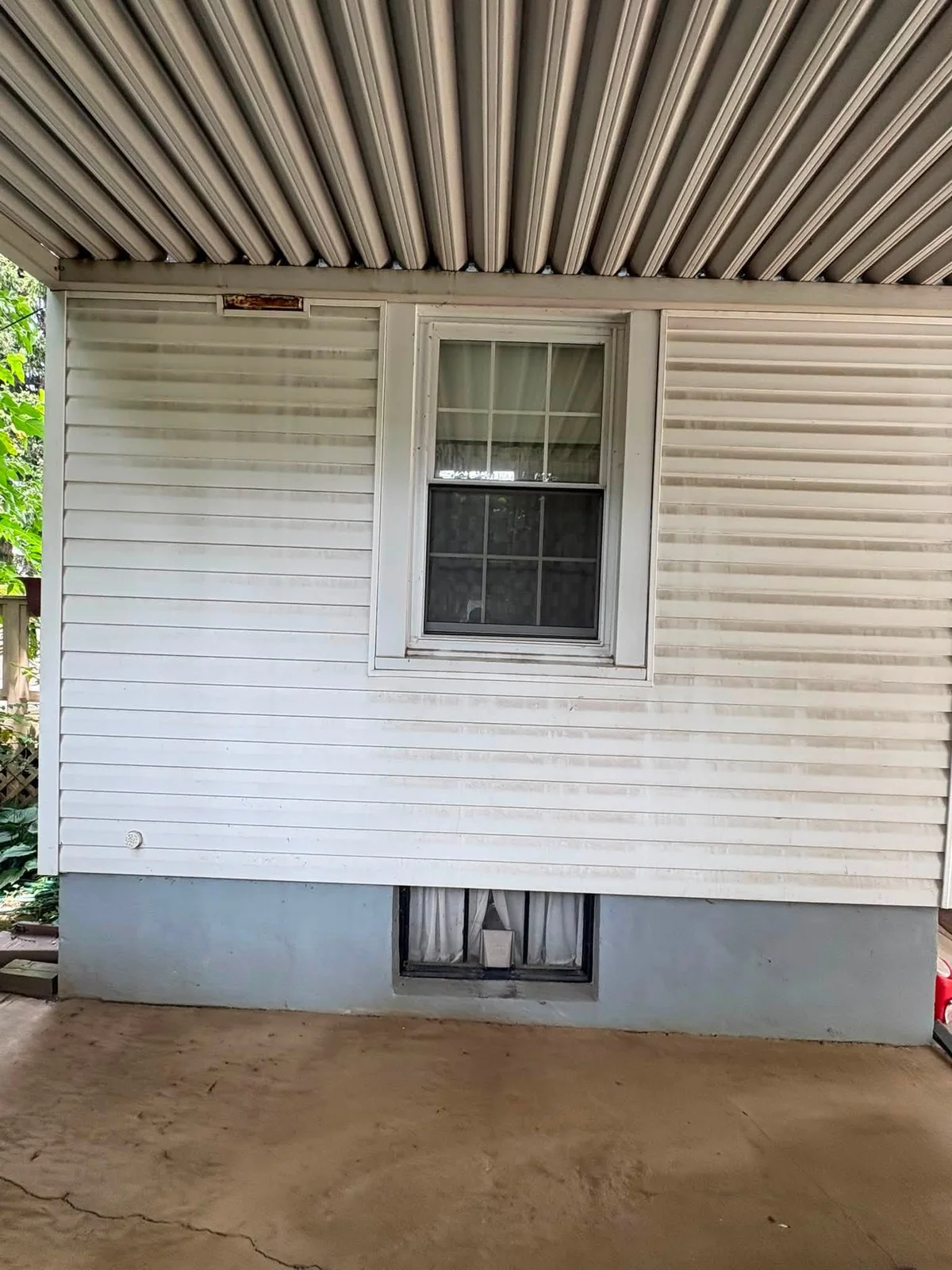 Before: ribbed metal carport ceiling over white vinyl siding with gray horizontal streaks, a window and a concrete floor