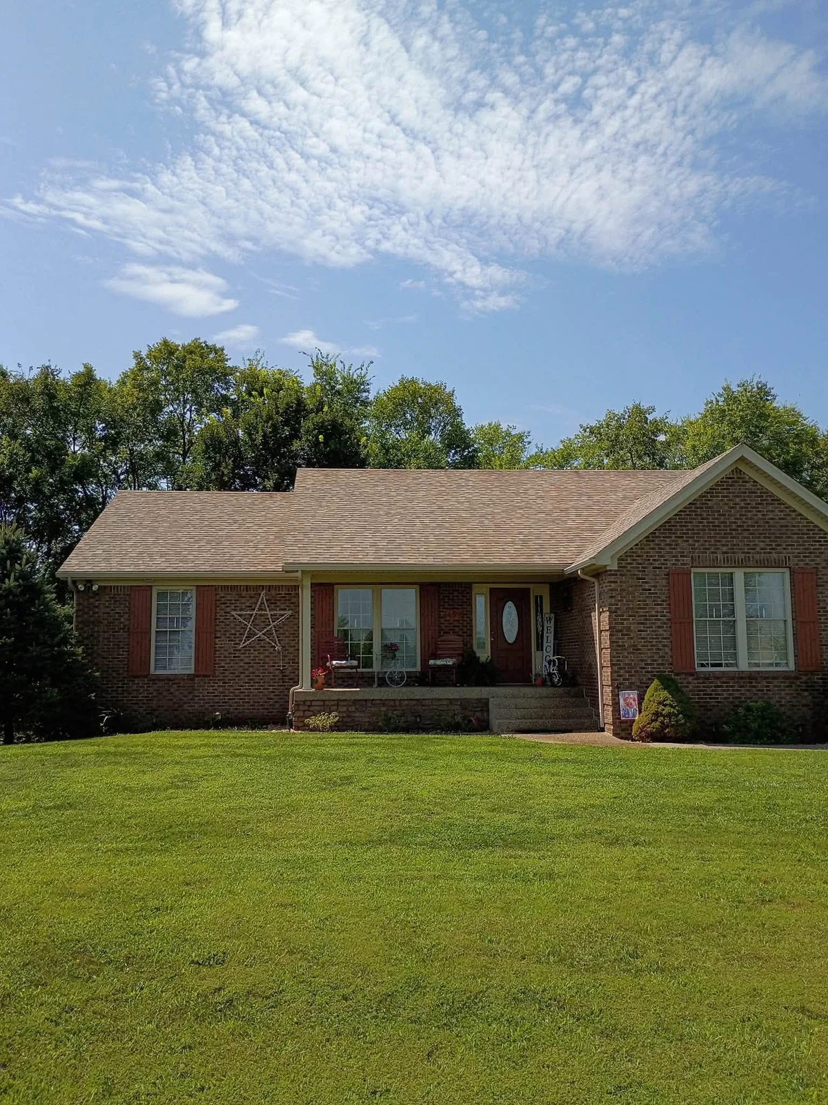 Single-story red brick ranch house with a tan shingle roof, red shutters, a covered front porch and a green lawn