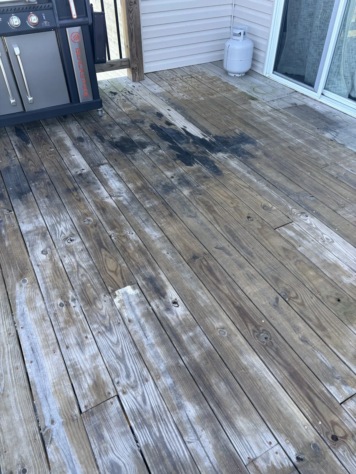 Before: gray weathered wood deck boards with dark stains and white patches, a gas grill at left and a propane tank by the sliding door