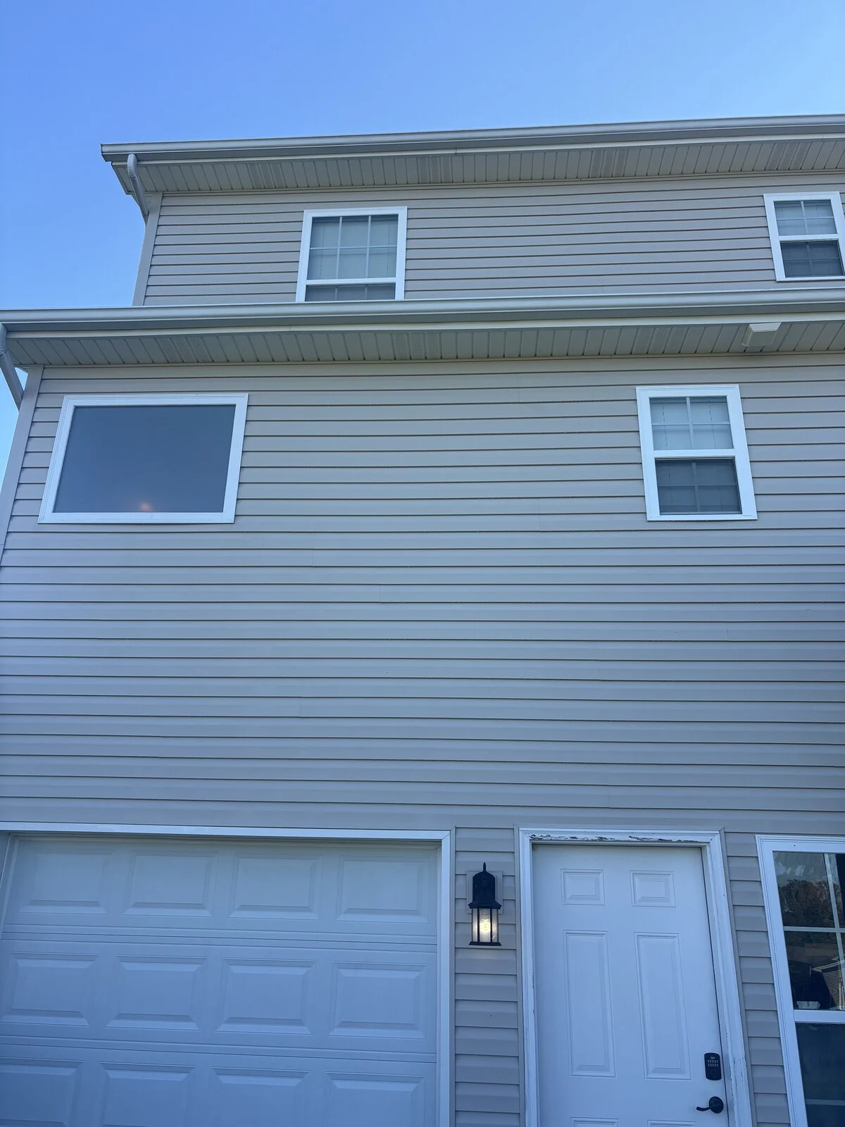 After: the same beige vinyl siding wall from farther back, with an even color, a white garage door, a white entry door and a wall lantern