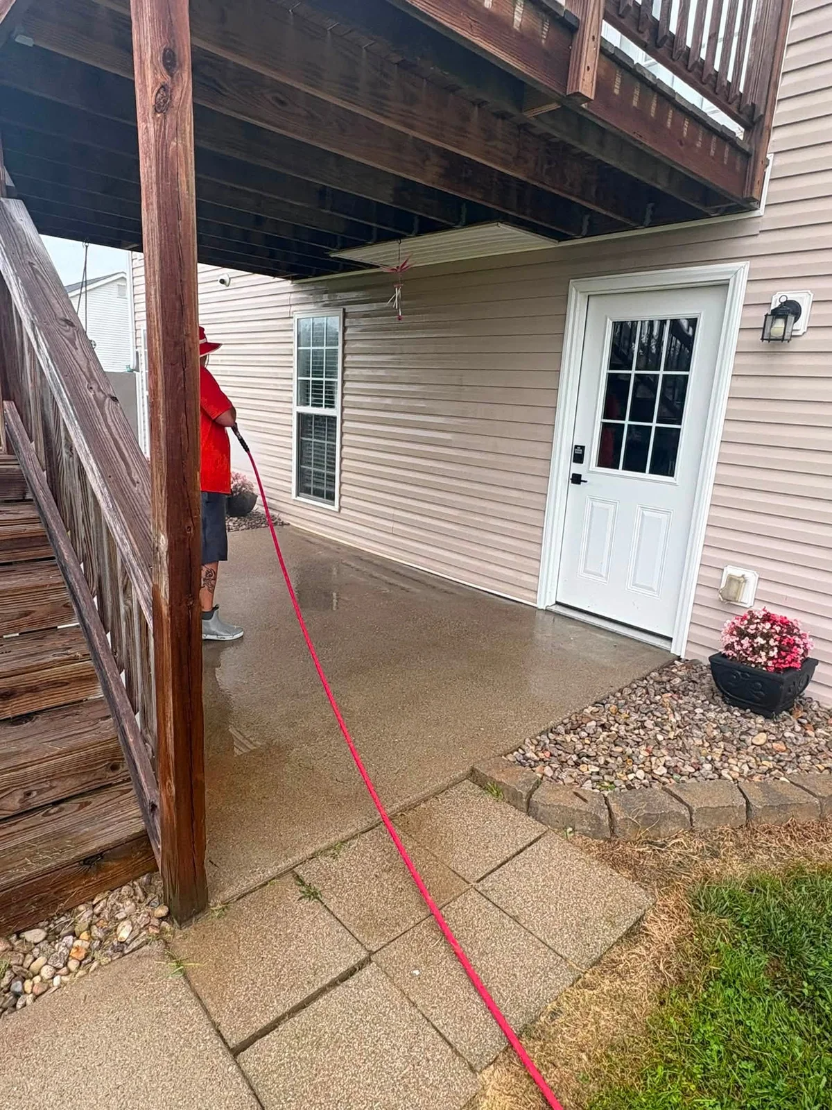 Crew member in a red shirt washing the concrete patio under a wood deck, red hose in hand, beside tan vinyl siding