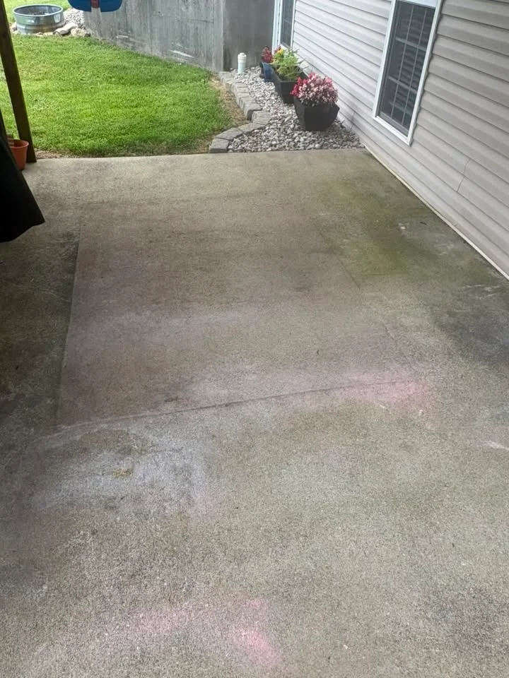Before: concrete patio with dark, green and gray stained patches beside tan vinyl siding, flower pots and a gravel bed